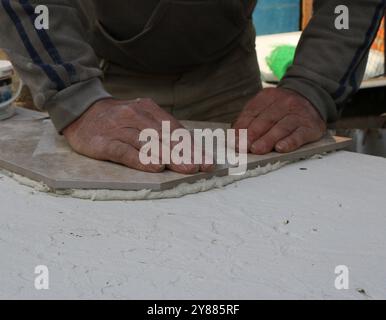 le carreleur dispose des morceaux de carreaux de céramique sur la surface du produit avec du mortier et met un poids de fer pour le niveler, face à une table ronde Banque D'Images