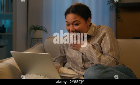 Riant femme afro-américaine souriante fille détendue repos féminin sur le canapé à la soirée de loisirs de nuit à la maison avec ordinateur portable regarder le film rire rire ayant Banque D'Images