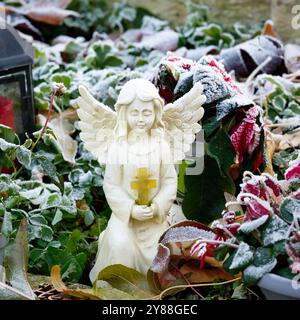 une petite figure blanche d'un ange avec une croix dans les mains pliées sur une tombe avec des plantes couvertes de givre Banque D'Images