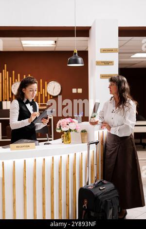 Une dame âgée se tient à la réception de l'hôtel, où un concierge professionnel vous aide à réserver. Femme senior complète le processus d'enregistrement avec document numérique sur tablette. La valise est placée près du comptoir. Banque D'Images