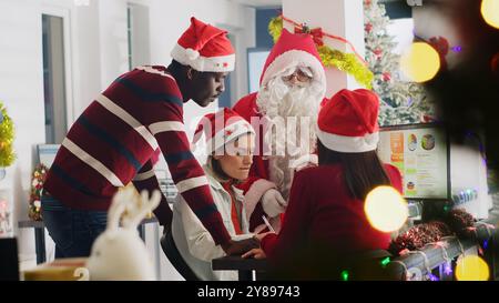 Le chef d'équipe habillé en Père Noël délègue les tâches du projet aux travailleurs. Directeur dans l'espace de travail décoré de Noël discutant avec les employés pendant la saison des vacances d'hiver Banque D'Images
