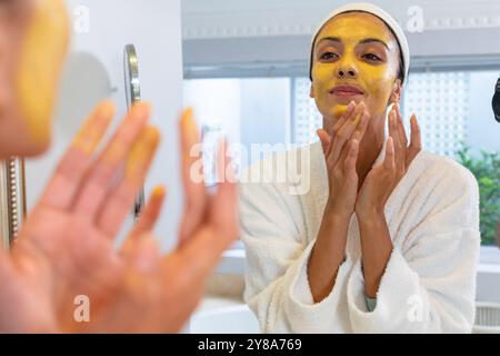 Jeune femme caucasienne regardant un miroir et appliquant un masque facial dans la salle de bain à la maison Banque D'Images