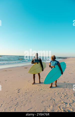 Couple aîné afro-américain avec planches de surf marchant à la plage de sable contre la mer et le ciel bleu Banque D'Images