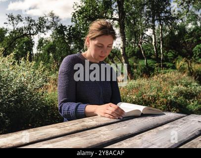 Femme assise à une table en bois, profondément imprégnée dans la lecture d'un livre entouré de verdure vibrante et d'arbres, profitant d'un moment de tranquillité dans la nature Banque D'Images