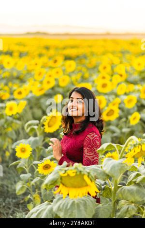 Femme dans une robe rouge debout dans un champ de tournesol, souriant au coucher du soleil. Banque D'Images