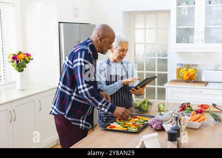 Couple senior diversifié préparant le repas en utilisant la tablette dans la cuisine Banque D'Images