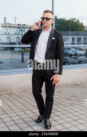 Homme élégant dans un costume noir avec des lunettes de soleil s'engageant sur un appel téléphonique dans un cadre urbain. Banque D'Images