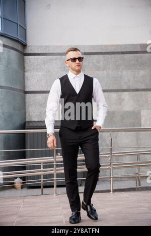 Homme élégant dans une tenue élégante avec des lunettes de soleil posant en toute confiance à l'extérieur. Banque D'Images