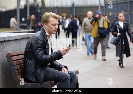 Gars dans des écouteurs assis avec smartphone sur le banc de rue en ville Banque D'Images