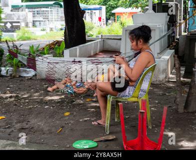 Manille, Philippines - 26 août 2019 : une mère est assise sur une chaise dans un cimetière de Manille, s'occupant de son enfant tandis qu'un autre enfant repose dans un hamac Banque D'Images