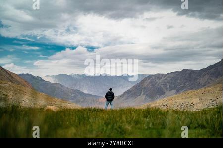 Une figure solitaire se tient sur des plaines herbeuses, regardant les majestueuses montagnes imposantes sous un ciel nuageux, embrassant la solitude. Banque D'Images