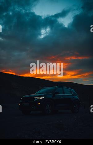 L'image montre un SUV garé contre un paysage accidenté au crépuscule. Un coucher de soleil dramatique aux teintes orange et rouge vives contraste avec des nuages bleu profond. Banque D'Images