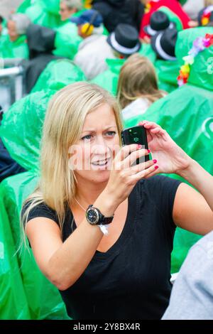 SMARTPHONE PRÉCOCE, TEMPÊTE ESTIVALE, BEACH VOLLEY, MARIEHAMN, 2011 : une femme blonde prend une photo avec un smartphone précoce. La foule se couvre avec des ponchos jetables alors qu'une énorme tempête estivale balaye le stade avec un déluge de pluie pendant les demi-finales en août 2011 au PAF Open à Mariehamn, Åland, Finlande. Photographie : Rob Watkins. INFO : entre 2009-2013, le tournoi PAF Open Beach Volleyball était un événement annuel organisé à Mariehamn, Åland, Finlande. Il a attiré les meilleures équipes et joueurs internationaux en tant que partie du classement du circuit mondial officiel de la FIVB, mettant en vedette le haut niveau Banque D'Images