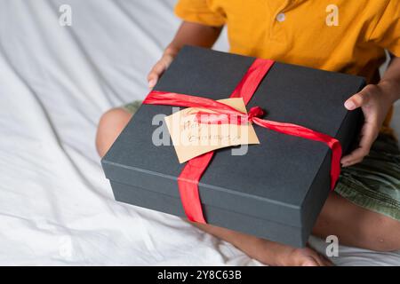 Un enfant tenant une boîte-cadeau noire magnifiquement enveloppée avec un ruban rouge et une étiquette qui dit joyeux Noël!a L'enfant est assis sur un lit blanc, porter Banque D'Images