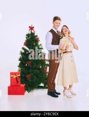 Couple pose gaiement devant le sapin de Noël, avec des cadeaux rouges en dessous, tenant des verres à champagne dans une atmosphère festive et élégante. Banque D'Images