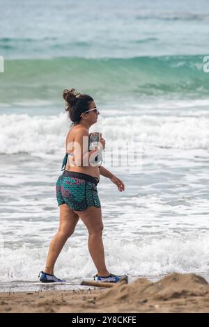 jeune femme légèrement en surpoids marchant le long d'une plage seule avec des vagues se brisant en arrière-plan, dame marchant sur une plage de sable au bord de l'eau. Banque D'Images