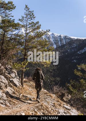 Un randonneur solitaire avec un sac à dos marche le long d'un étroit sentier de montagne dans les Alpes françaises. Les sommets enneigés et les forêts denses en arrière-plan créent Banque D'Images