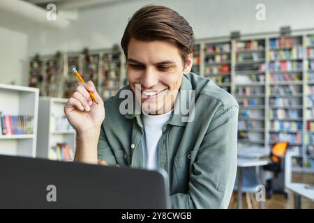 Un jeune homme joyeux étudie avec diligence dans une bibliothèque bien éclairée, entourée d'étagères de livres. Banque D'Images