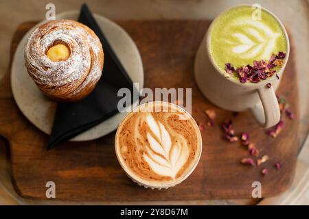 Plateau en bois avec cappuccino et latte matcha avec latte art, accompagné d'un croissant en poudre. Cadre confortable de café avec décor rustique. Banque D'Images