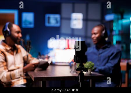 L'animateur du podcast utilise un smartphone sur un trépied pour enregistrer les invités donnant aux fans des histoires fascinantes depuis le salon. Invité au home studio, la célébrité masculine partage des faits avec l'intervieweur. Banque D'Images