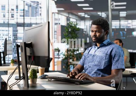 Employé de démarrage assis au bureau dans le bureau, faisant des recherches pour le projet avec la date limite approchant. Homme afro-américain regardant l'ordinateur dans l'espace de travail, résolvant des tâches pendant le quart de travail Banque D'Images