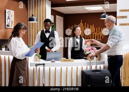 Une dame âgée au comptoir remplit les documents de réservation tandis que la réceptionniste aimable conseille un vieil homme dans le hall de l'hôtel. Les employés professionnels fournissent un bon service à la clientèle aux clients. Banque D'Images