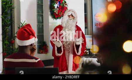 Le gérant s'habillait en Père Noël pendant la période de Noël, organisant une réunion d'équipe et présentant des graphiques commerciaux sur un écran numérique. Chef d'équipe dans le bureau décoré festif discutant avec les membres du personnel Banque D'Images