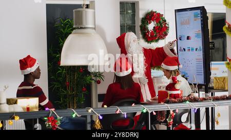 Le gérant s'habillait en Père Noël pendant la période de Noël, organisant une réunion d'équipe et présentant des graphiques financiers sur un écran numérique. Chef d'équipe dans le bureau décoré festif motiver les travailleurs Banque D'Images