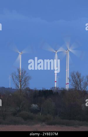 Trois éoliennes debout dans un paysage d'arbres au crépuscule, éoliennes dans la lumière du soir, longue exposition, parc naturel Flusslandschaft Peenetal Banque D'Images