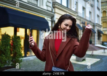 Belle jeune femme brune joyée aux cheveux longs dans un manteau rouge célébrant les bonnes nouvelles tenant le téléphone et serrant poing dans la rue du soir Banque D'Images