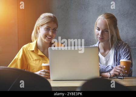 Deux jolies filles en vêtements décontractés regardent l'écran d'un ordinateur portable tout en étant assis au bureau. Les blogueurs ou les concepteurs lisent des nouvelles sur un ordinateur portable qui Banque D'Images