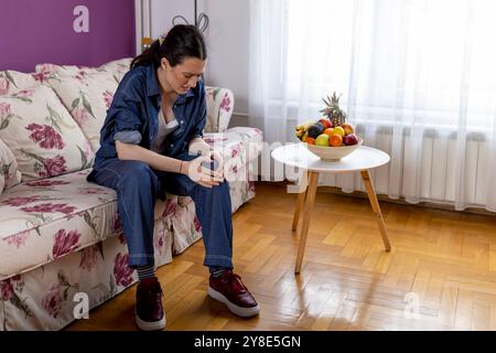 Une femme, elle a mal au genou sur le canapé dans le salon à la maison, la santé et le concept de style de vie Banque D'Images