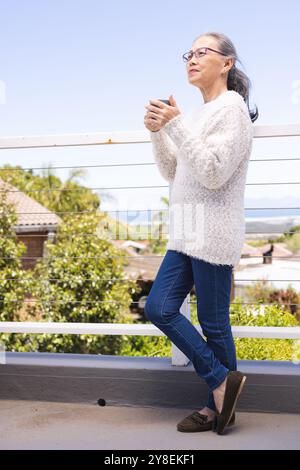 Heureuse femme asiatique senior tenant une tasse et regardant loin sur balkony Banque D'Images