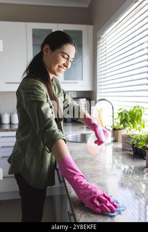 Vue latérale d'une jeune femme caucasienne portant des gants de vaisselle roses nettoyant le comptoir de cuisine Banque D'Images