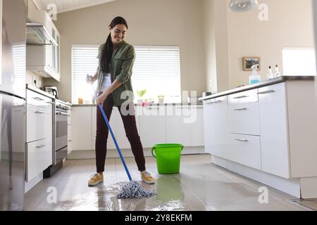 Jeune femme souriante caucasienne nettoyant le sol de la cuisine avec vadrouille humide à la maison, espace de copie Banque D'Images