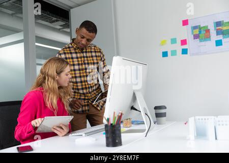 Vue latérale d'un young African American man standing et d'une jeune femme de race blanche assis à un bureau à l'aide d'ordinateurs tablettes dans les bureaux modernes d'un crea Banque D'Images