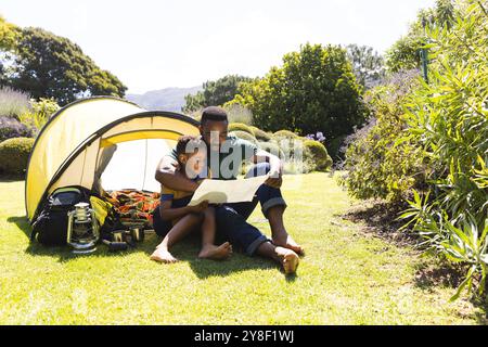 Heureux père afro-américain et fils assis sur l'herbe et lisant la carte à côté de tente dans le jardin Banque D'Images