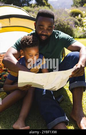 Heureux père afro-américain et fils assis sur l'herbe et lisant la carte à côté de tente dans le jardin Banque D'Images