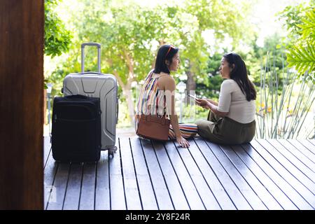 Heureuses amies asiatiques avec des bagages assis et parlant dans le jardin à la maison Banque D'Images