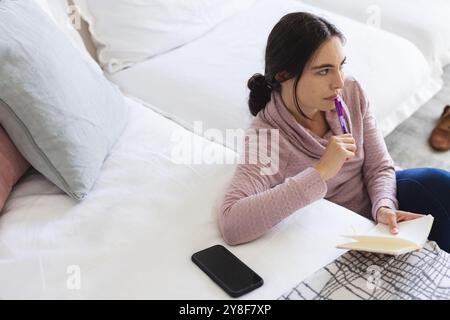 Vue en angle élevé de la jeune femme caucasienne pensant et écrivant dans le bloc-notes tout en se relaxant par canapé Banque D'Images