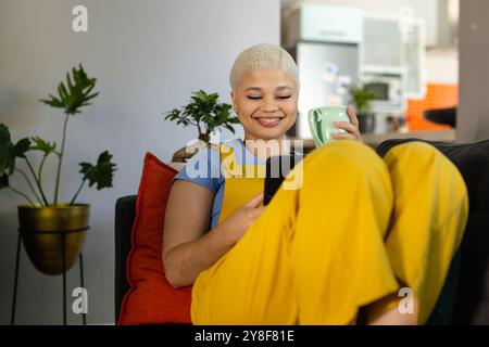 Heureuse femme biraciale inaltérée à la mode se relaxant sur le canapé à la maison avec du café, à l'aide d'un smartphone Banque D'Images