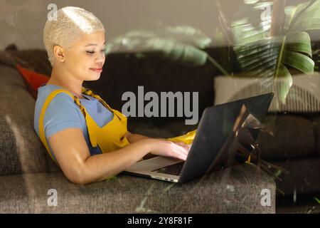 Détendu femme biraciale inaltérée à la mode assise sur le canapé à la maison à l'aide d'un ordinateur portable et souriant Banque D'Images