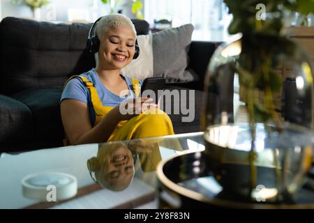 Heureuse femme biraciale inaltérée à la mode assise sur le sol à la maison dans des écouteurs à l'aide d'un smartphone Banque D'Images