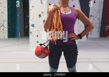 Section médiane de femme caucasienne avec casque et corde se préparant à grimper au mur d'escalade intérieur Banque D'Images