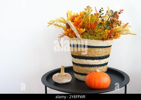 Panier tissé rempli de fleurs d'automne colorées sur une table noire avec une petite citrouille sur fond de mur blanc, améliorant l'ambiance saisonnière du ro Banque D'Images