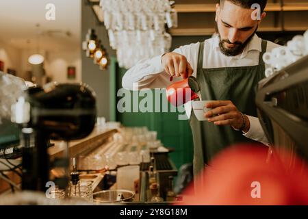 Dans un café chaleureux et accueillant, un barista dévoué verse habilement du lait dans une tasse à café, créant une expérience délicieuse pour les clients. Banque D'Images