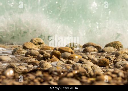 Cette image capture une vue rapprochée de galets lisses et arrondis sur une plage avec une vague s'écrasant en arrière-plan. Banque D'Images