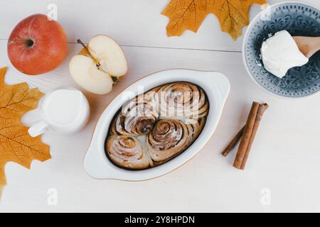 Des petits pains à la cannelle fraîchement cuits reposent dans un plat à côté de pommes tranchées, d'un verre de lait et d'un bol de crème glacée, entourés de feuilles d'automne. Banque D'Images
