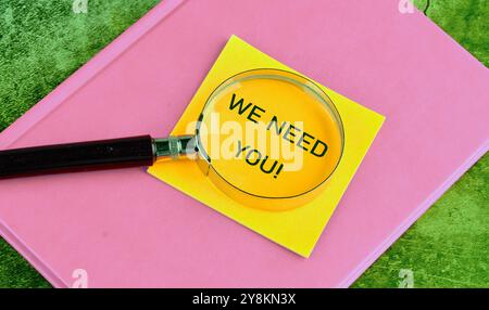 Nous avons besoin de votre symbole. Mots conceptuels NOUS AVONS BESOIN DE VOUS sur un autocollant jaune à travers une loupe couchée sur un carnet rose Banque D'Images