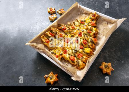 Arbre de Noël de vacances cuit au four et étoiles de pâte feuilletée avec pesto, tomate et graines, servi sur un plateau avec du papier sulfurisé, fond d'ardoise foncé, créat Banque D'Images